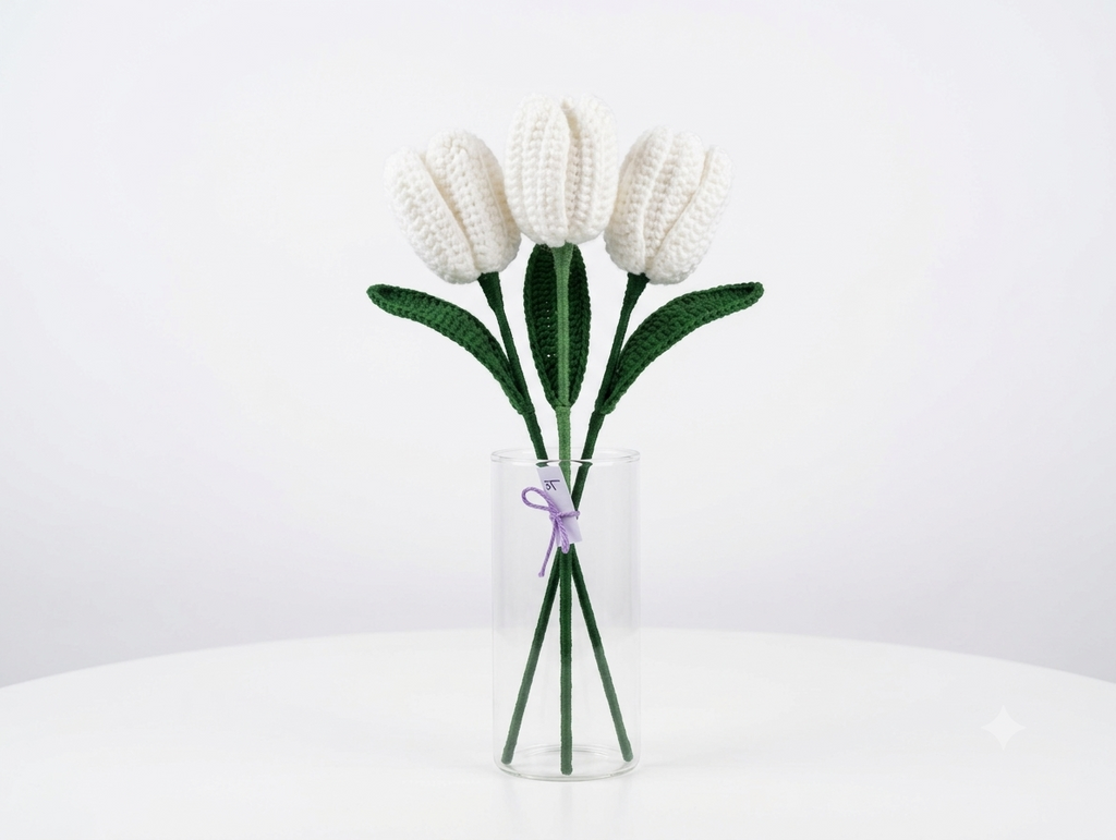Three white crochet tulips with green leaves in a tall glass vase against a white background