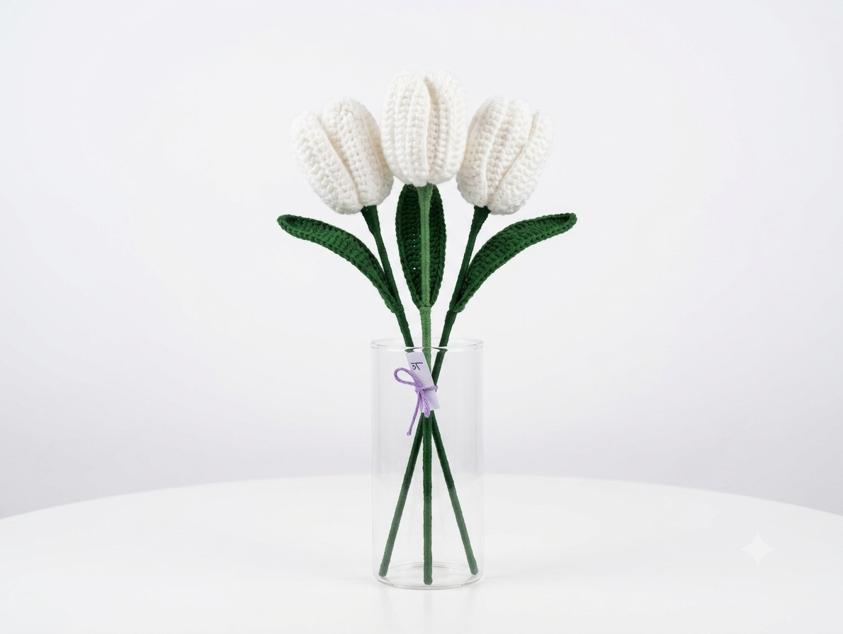 Three white crochet tulips with green leaves in a tall glass vase against a white background
