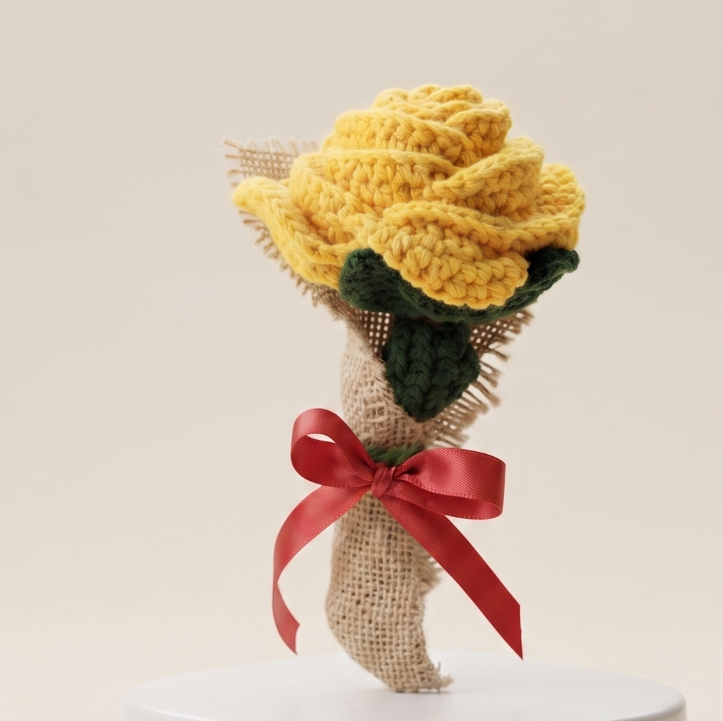 Handcrafted chunky crochet yellow rose with green leaves wrapped in burlap and red satin bow on white pedestal