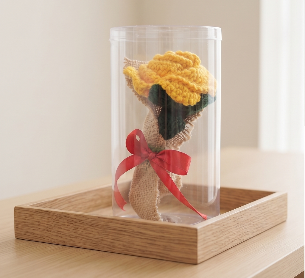Handcrafted crochet yellow rose in burlap with red bow displayed inside clear cylindrical gift box on wooden tray