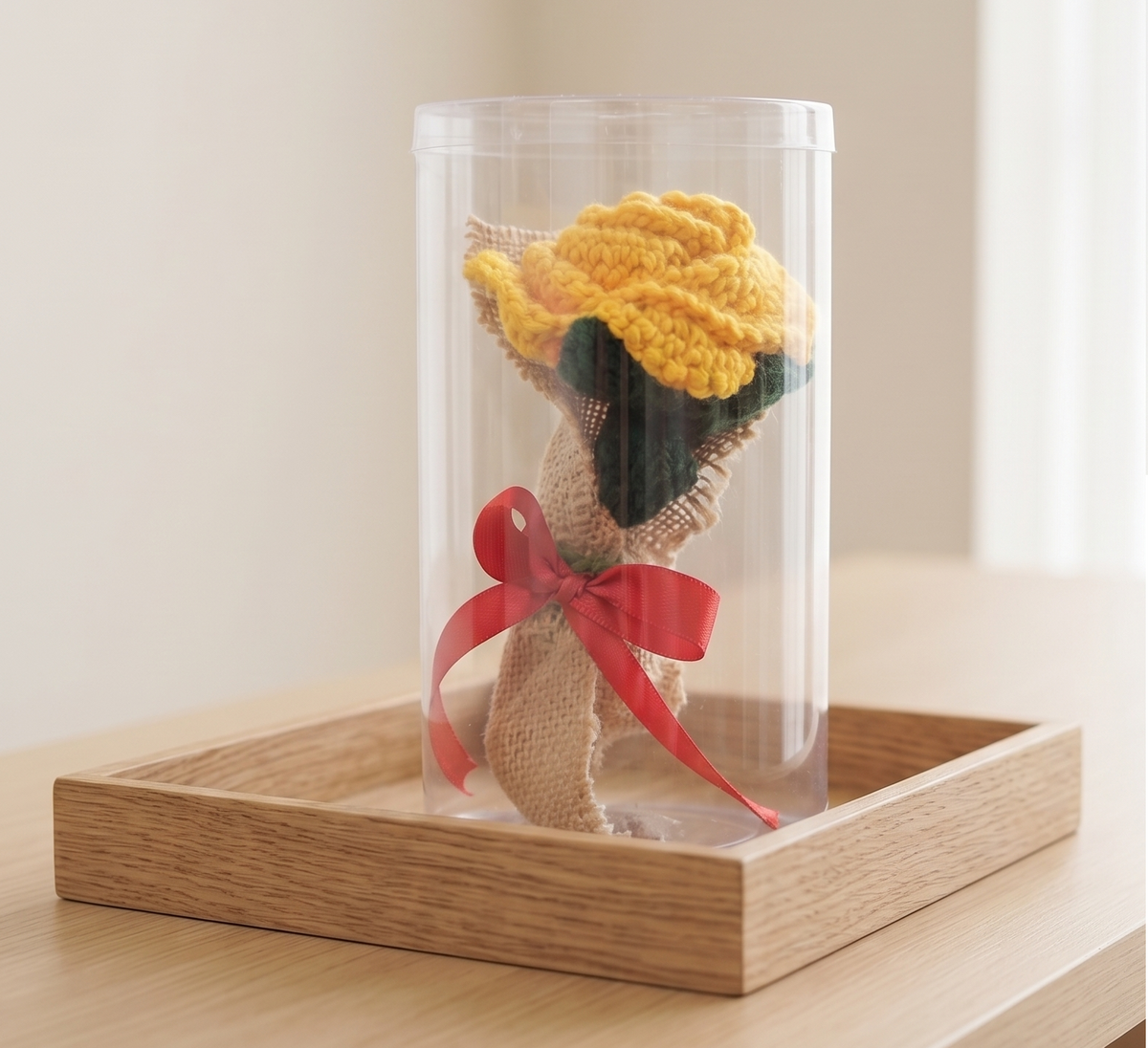 Handcrafted crochet yellow rose in burlap with red bow displayed inside clear cylindrical gift box on wooden tray